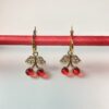 Aros Colgantes Dije Cereza Dorados Par de aros colgantes de acero quirúrgico dorado con dije de cerezas rojas y hojas con cristales, mostrados sobre una base roja.