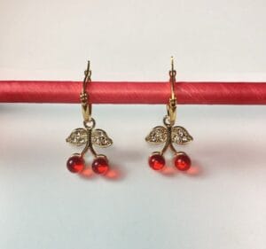 Aros Colgantes Dije Cereza Dorados Par de aros colgantes de acero quirúrgico dorado con dije de cerezas rojas y hojas con cristales, mostrados sobre una base roja.