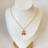 Collar Cereza Dorado en Exhibidor Collar de acero dorado con dije de cerezas rojas y hojas con cristales, exhibido en un busto de joyería blanco sobre base de madera.