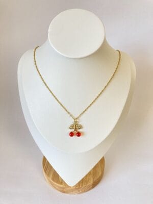 Collar Cereza Dorado en Exhibidor Collar de acero dorado con dije de cerezas rojas y hojas con cristales, exhibido en un busto de joyería blanco sobre base de madera.
