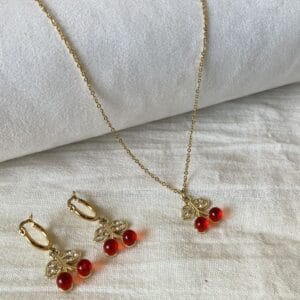 Portada Set Collar y Aros Cereza Set de joyas para mujer de acero dorado hipoalergénico, mostrando el collar y los aros con dijes de cereza sobre una tela de lino blanco.