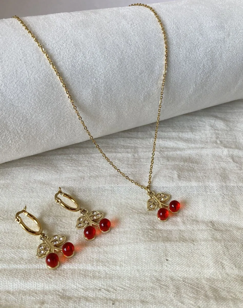 Set de joyas para mujer de acero dorado hipoalergénico, mostrando el collar y los aros con dijes de cereza sobre una tela de lino blanco.
