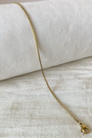 Collar Fino de Acero Dorado Modelo Cuadros Collar de acero dorado con cadena de eslabones cuadrados (box chain), conocido como modelo Cuadros, sobre una tela de lino blanco.