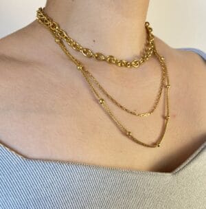 Set de Collares en Capas (Layering) en Modelo Vista lateral de una modelo luciendo un set de tres collares de acero dorado en capas: un choker de eslabones gruesos, una cadena fina rectangular y una cadena de bolitas.