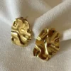 Detalle Aros Chunky Dorados Par de aros dorados de acero quirúrgico con textura irregular sobre una tela blanca.