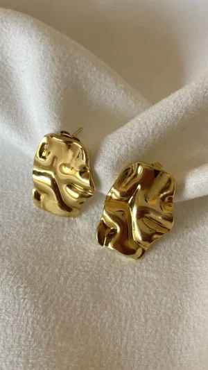 Detalle Aros Chunky Dorados Par de aros dorados de acero quirúrgico con textura irregular sobre una tela blanca.
