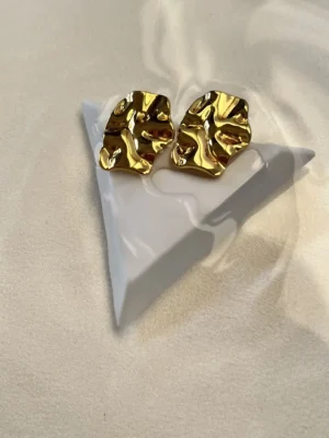 Diseño Artístico Aros Dorados Aros chunky dorados con diseño molten sobre una figura geométrica blanca.