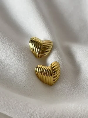 Aros Corazón Acero Quirúrgico Par de aros de corazón dorados de acero quirúrgico sobre tela blanca.