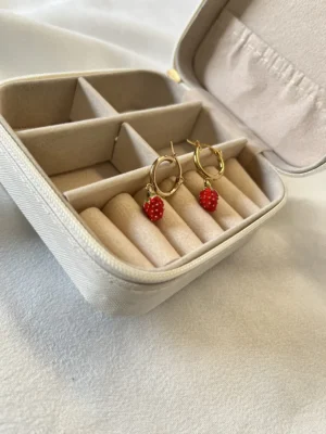 Aros Coquette en Alhajero Aros de argollita con dije de frutilla guardados en un alhajero de viaje color crema.