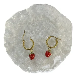 Detalle Aros Frutilla Esmaltados Par de aros de argolla con dije de frutilla esmaltada sobre una base de vidrio texturizada.