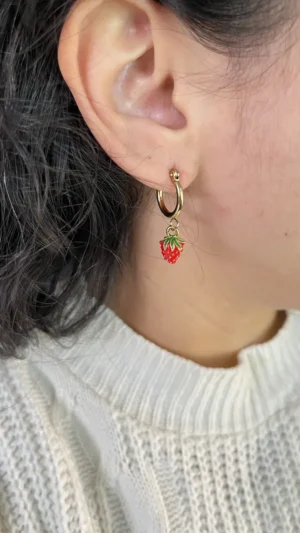 Aros Frutilla Coquette en Oreja Primer plano de una mujer usando un aro de argollita dorada con un dije de frutilla roja.
