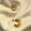 Diseño Entrelazado Aros Triple Argolla Par de aros de triple argolla dorada entrelazados sobre una superficie blanca.