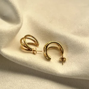 Aros Triple Argolla Dorados Par de aros de triple argolla dorada de acero quirúrgico sobre una tela blanca.