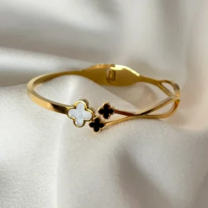 Pulsera Dorada Trébol de la Suerte Pulsera esclava dorada con un trébol de nácar blanco y dos tréboles negros sobre una tela blanca.